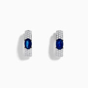 The Royal Bleu 14K White Gold Sapphire and Diamond Huggie Earrings showcase oval sapphire centers surrounded by rows of sparkling diamonds, displayed against a plain white background.