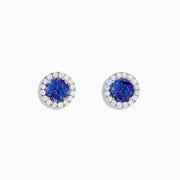 The Nahla 14K White Gold Tanzanite and Diamond Stud Earrings feature round deep blue tanzanite centers, each surrounded by a halo of sparkling white diamonds, beautifully set in 14K white gold against a crisp white background.
