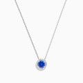 The Nahla 14K White Gold Tanzanite and Diamond Halo Necklace features a round blue tanzanite pendant encircled by sparkling diamonds on a delicate silver chain, creating an elegant look against a plain white background.