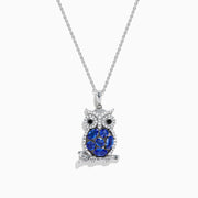The Nahla 925 Silver Tanzanite and Black Diamond Owl Pendant features black diamond eyes and sparkling tanzanite gemstones on its body, perched on a branch against a white background.