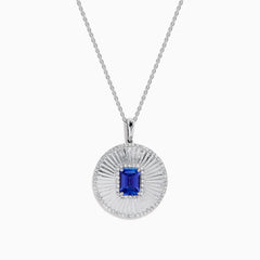 The 14K White Gold Tanzanite and Diamond Fluted Circle Pendant features a round diamond-accented design with a radiant motif and a central emerald-cut blue tanzanite stone.