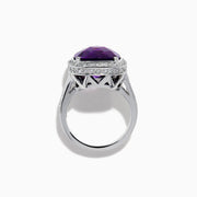 The 18K White Gold Amethyst and Diamond Cushion Halo Cocktail Ring features a striking cushion-cut amethyst center, a sparkling diamond halo, and clear stones on the band, elegantly displayed against a white background.