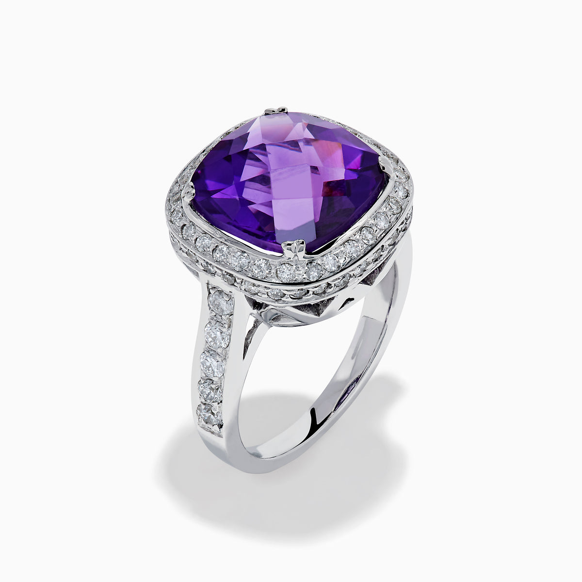 The 18K White Gold Amethyst and Diamond Cushion Halo Cocktail Ring features a striking cushion-cut amethyst center, a sparkling diamond halo, and clear stones on the band, elegantly displayed against a white background.