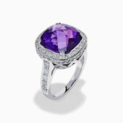 The 18K White Gold Amethyst and Diamond Cushion Halo Cocktail Ring features a striking cushion-cut amethyst center, a sparkling diamond halo, and clear stones on the band, elegantly displayed against a white background.