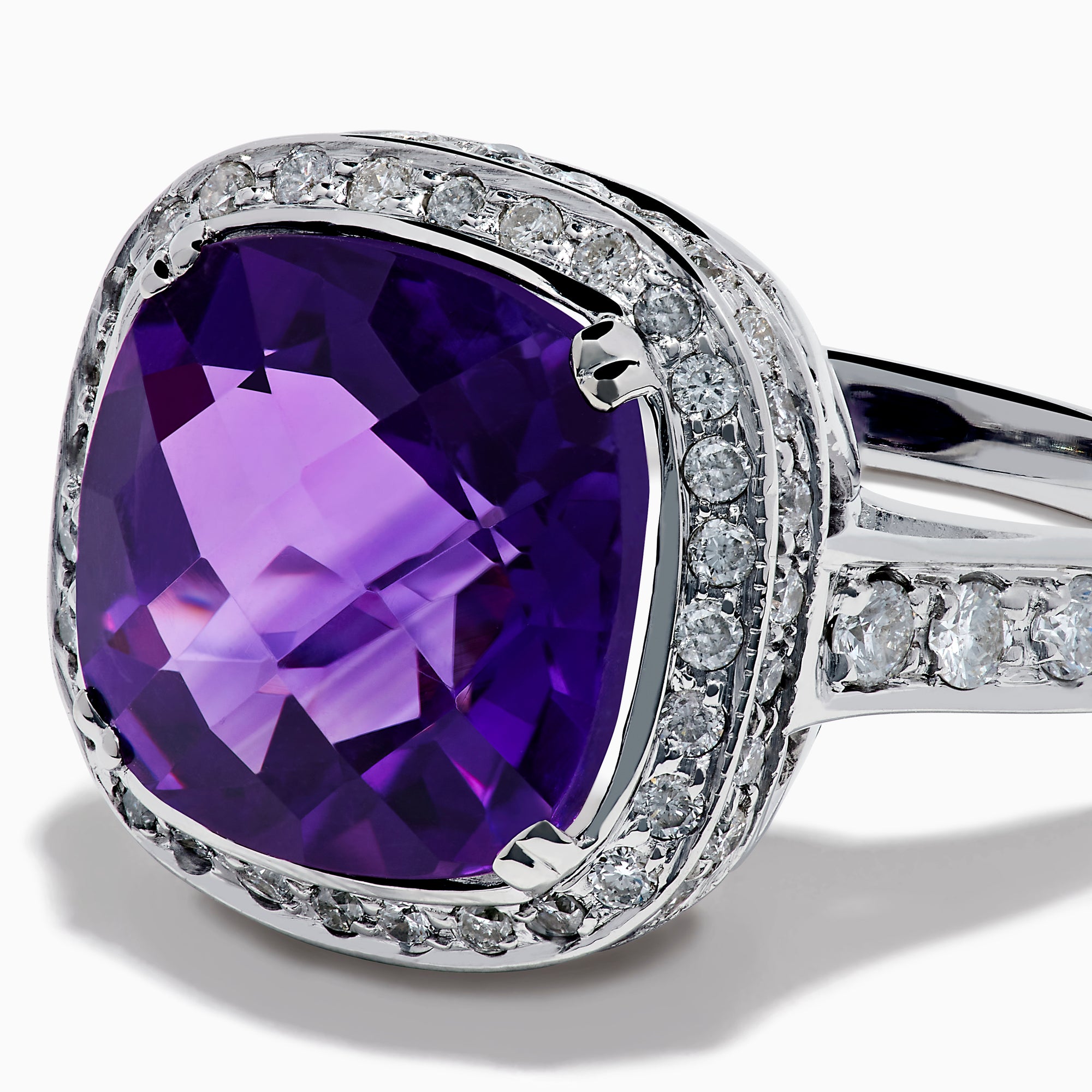 The 18K White Gold Amethyst and Diamond Cushion Halo Cocktail Ring features a striking cushion-cut amethyst center, a sparkling diamond halo, and clear stones on the band, elegantly displayed against a white background.