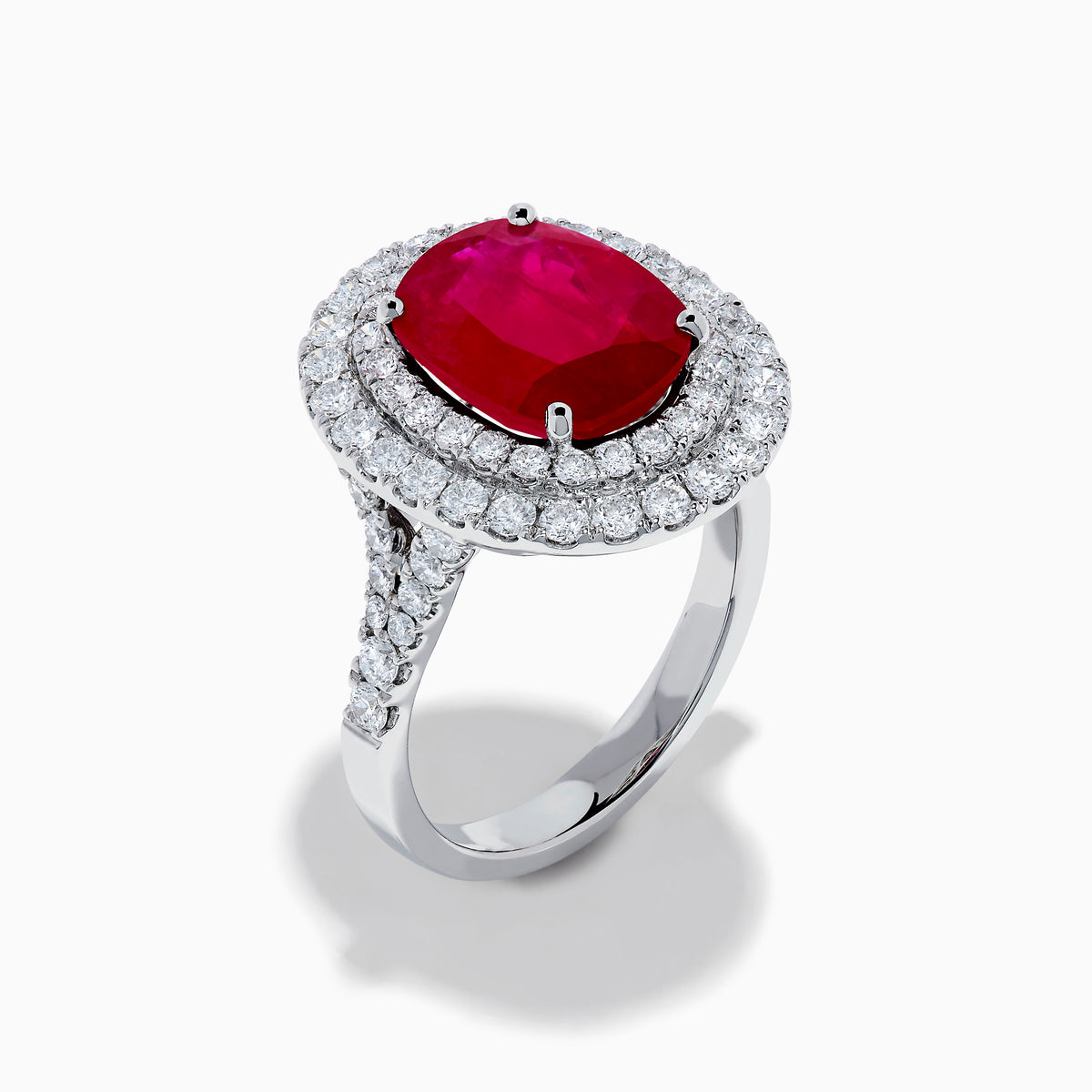 The 18K White Gold Ruby and Diamond Double Halo Ring features an oval ruby encircled by two rows of round white diamonds, set in a sleek 18K white gold band and displayed on a white background.