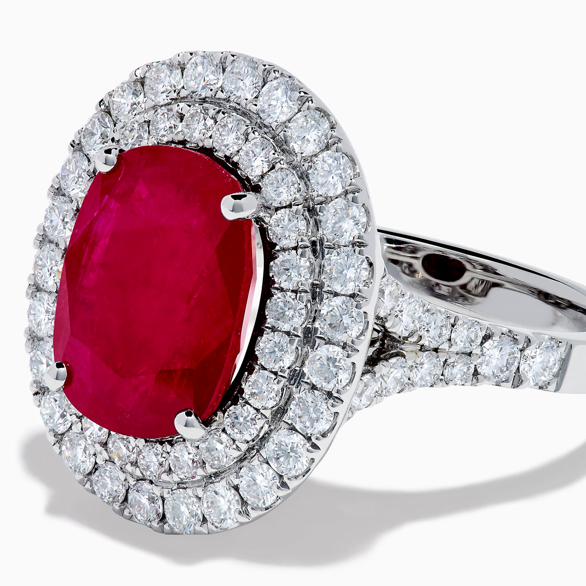 The 18K White Gold Ruby and Diamond Double Halo Ring features an oval ruby encircled by two rows of round white diamonds, set in a sleek 18K white gold band and displayed on a white background.