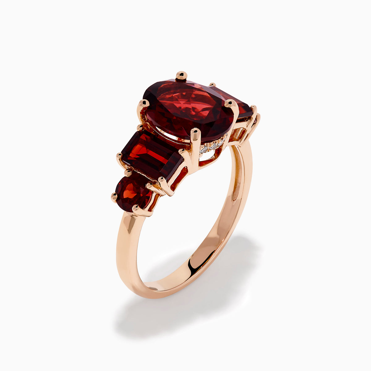The 14K Rose Gold Multi Shape Garnet and Diamond Hidden Halo Ring showcases a central oval deep red garnet, accented by smaller rectangular and oval garnets on each side, all set in polished rose gold.