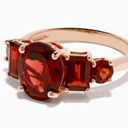 The 14K Rose Gold Multi Shape Garnet and Diamond Hidden Halo Ring showcases a central oval deep red garnet, accented by smaller rectangular and oval garnets on each side, all set in polished rose gold.