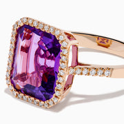 The 14K Rose Gold Amethyst and Diamond Halo Ring features a large, emerald-cut purple amethyst at the center, encircled by sparkling diamonds, and is showcased on a plain white background.