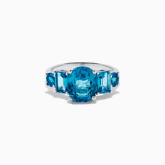 The 14K White Gold Multi Shape Blue Topaz and Diamond Hidden Halo Ring showcases a bold oval blue topaz center, accented by rectangular and round blue stones on each side, all set in a polished band with a subtle diamond hidden halo.