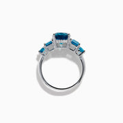 The 14K White Gold Multi Shape Blue Topaz and Diamond Hidden Halo Ring showcases a bold oval blue topaz center, accented by rectangular and round blue stones on each side, all set in a polished band with a subtle diamond hidden halo.
