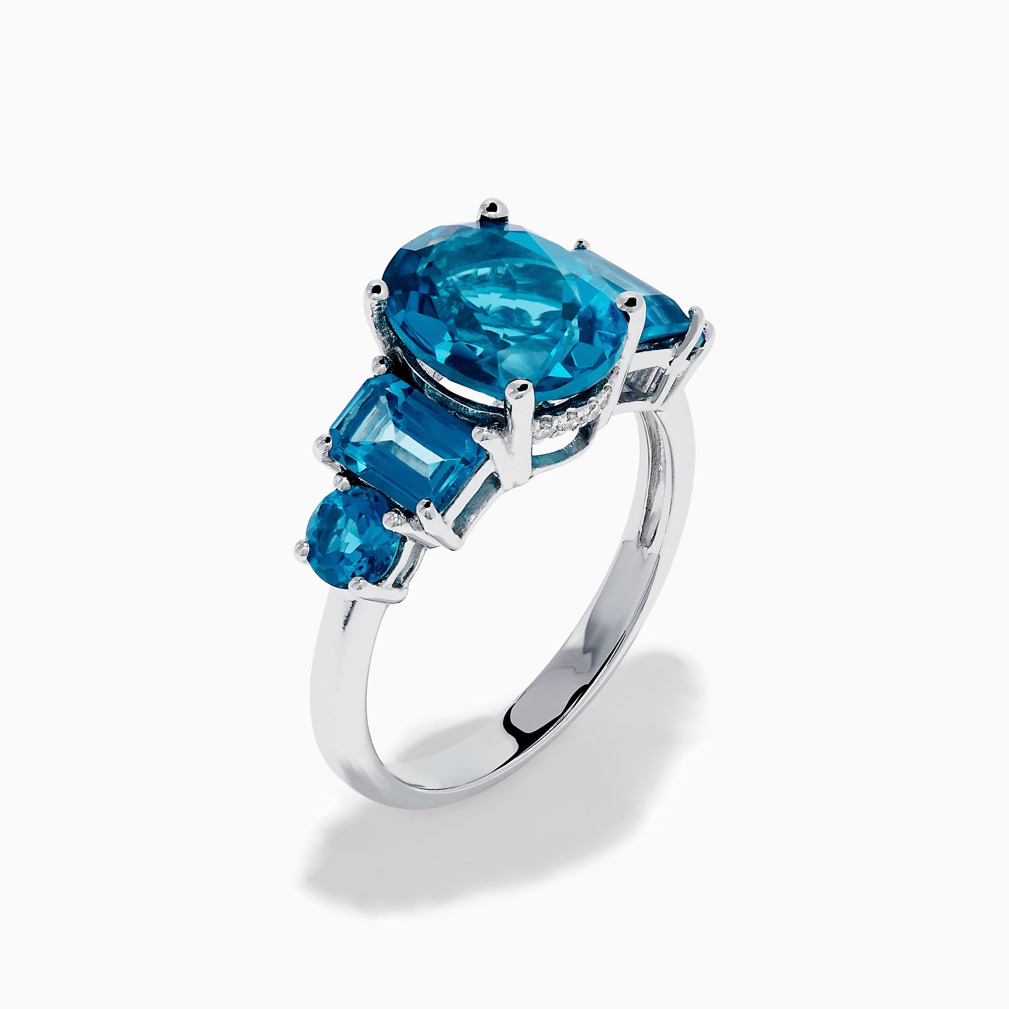 The 14K White Gold Multi Shape Blue Topaz and Diamond Hidden Halo Ring showcases a bold oval blue topaz center, accented by rectangular and round blue stones on each side, all set in a polished band with a subtle diamond hidden halo.