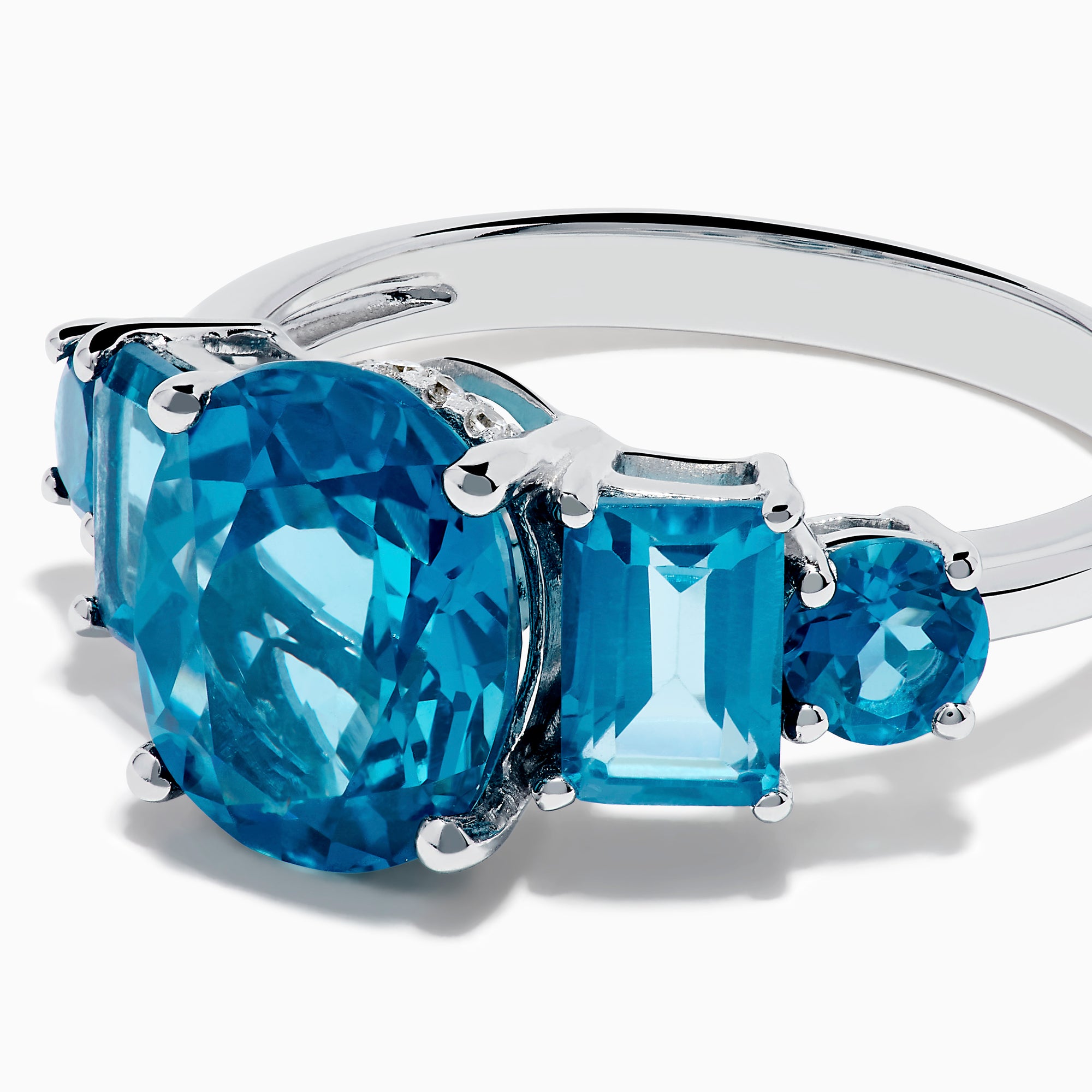 The 14K White Gold Multi Shape Blue Topaz and Diamond Hidden Halo Ring showcases a bold oval blue topaz center, accented by rectangular and round blue stones on each side, all set in a polished band with a subtle diamond hidden halo.