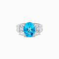 The Ocean Bleu 14K White Gold Blue Topaz and Diamond ring showcases a large oval blue topaz center stone, accented by two rectangular diamonds on each side, set against a white background.