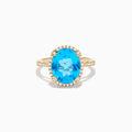 The Ocean Bleu 14K Yellow Gold Blue Topaz Ring showcases a large oval center blue topaz, embraced by a diamond halo and additional diamonds adorning the band.