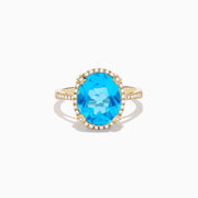 The Ocean Bleu 14K Yellow Gold Blue Topaz Ring showcases a large oval center blue topaz, embraced by a diamond halo and additional diamonds adorning the band.