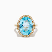 The 14K Yellow Gold Blue Topaz and Diamond Halo Cocktail Ring showcases a large oval blue topaz gemstone surrounded by sparkling diamonds, presented against a simple white background.