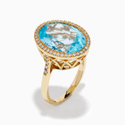 The 14K Yellow Gold Blue Topaz and Diamond Halo Cocktail Ring showcases a large oval blue topaz gemstone surrounded by sparkling diamonds, presented against a simple white background.
