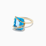 The Ocean Bleu 14K Yellow Gold Blue Topaz and Diamond Ring showcases a large emerald-cut blue topaz with two twisted gold bands—one plain, one adorned with diamonds—crisscrossing over the stone.