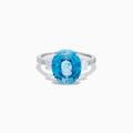 The 14K White Gold Blue Zircon and Diamond 3 Stone Ring features a large oval blue zircon center stone with diamonds on each side, shown on a white background.