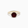 The 14K Yellow Gold Garnet and Diamond Ring features a large round deep red garnet center, accented by small white diamonds on each side of the band, displayed against a white background.