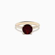 The 14K Yellow Gold Garnet and Diamond Ring features a large round deep red garnet center, accented by small white diamonds on each side of the band, displayed against a white background.