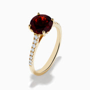 The 14K Yellow Gold Garnet and Diamond Ring features a large round deep red garnet center, accented by small white diamonds on each side of the band, displayed against a white background.