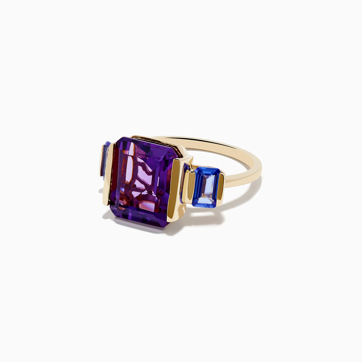 The 14K Yellow Gold Amethyst and Tanzanite 3 Stone Ring features a central emerald-cut amethyst with two rectangular tanzanites on each side, all set against a white background.