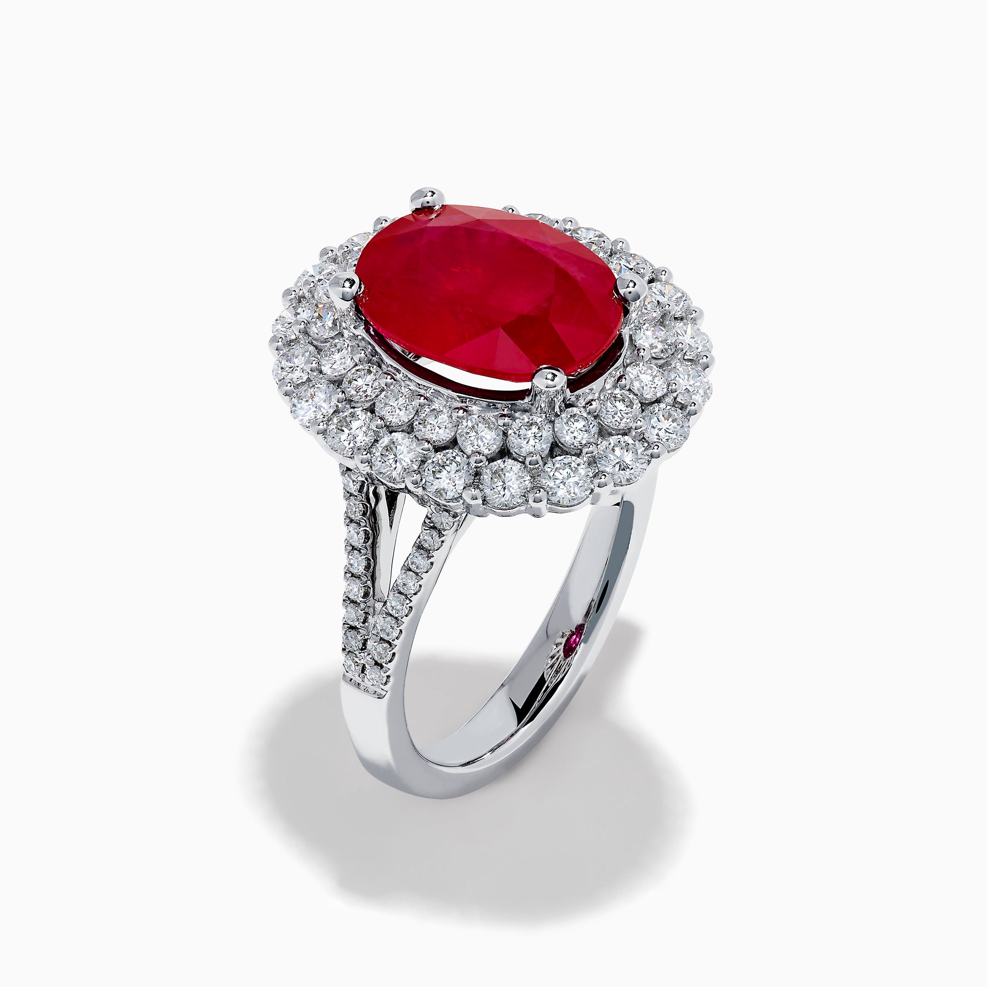 The 18K White Gold Ruby and Diamond Double Halo Ring features an oval ruby encircled by double diamond halos on a sleek white gold band, showcased against a white background.