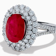 The 18K White Gold Ruby and Diamond Double Halo Ring features an oval ruby encircled by double diamond halos on a sleek white gold band, showcased against a white background.