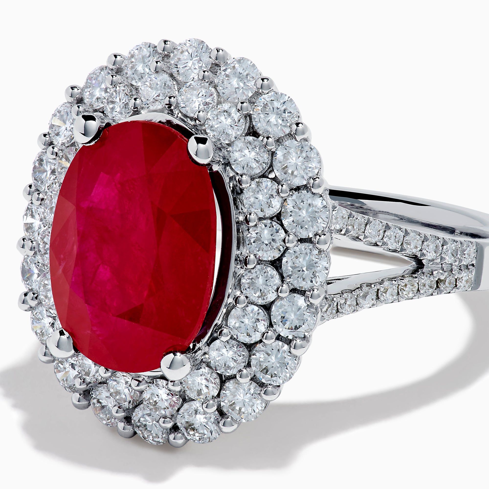 The 18K White Gold Ruby and Diamond Double Halo Ring features an oval ruby encircled by double diamond halos on a sleek white gold band, showcased against a white background.