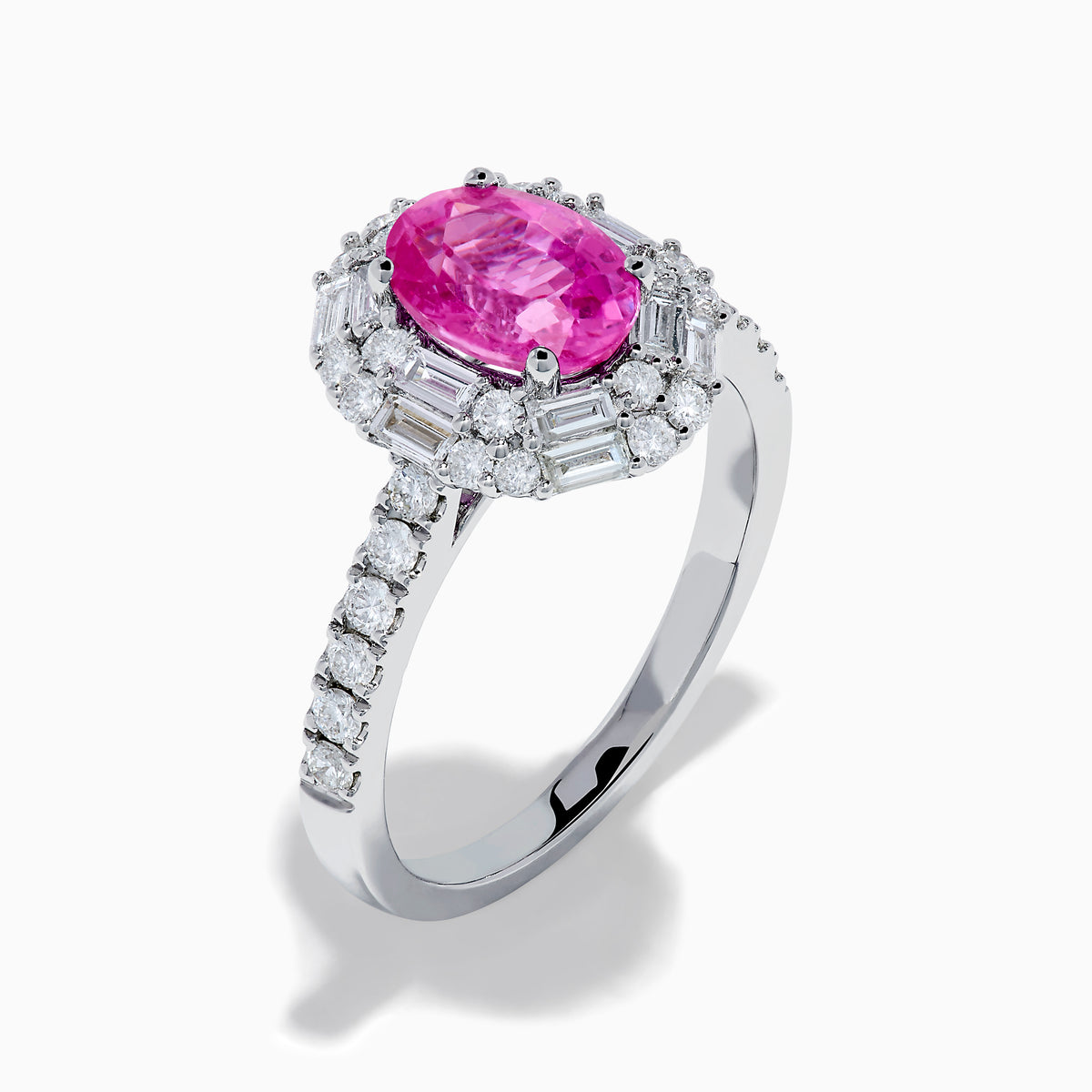 The 18K White Gold Pink Sapphire and Diamond Halo Ring showcases an oval pink sapphire center, encircled by a halo of baguette and round-cut diamonds, with more diamonds adorning the band.