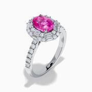 The 18K White Gold Pink Sapphire and Diamond Halo Ring showcases an oval pink sapphire center, encircled by a halo of baguette and round-cut diamonds, with more diamonds adorning the band.
