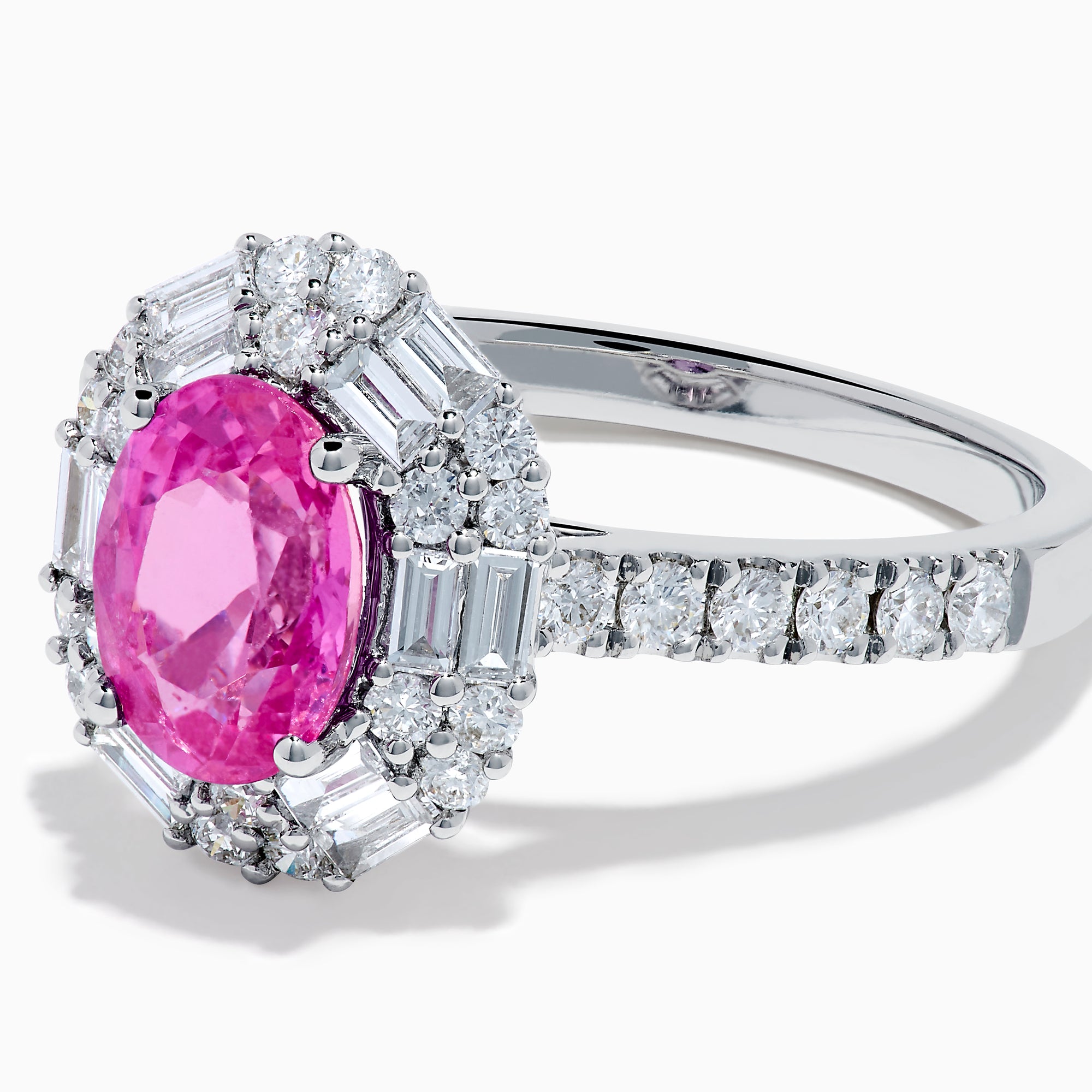 The 18K White Gold Pink Sapphire and Diamond Halo Ring showcases an oval pink sapphire center, encircled by a halo of baguette and round-cut diamonds, with more diamonds adorning the band.