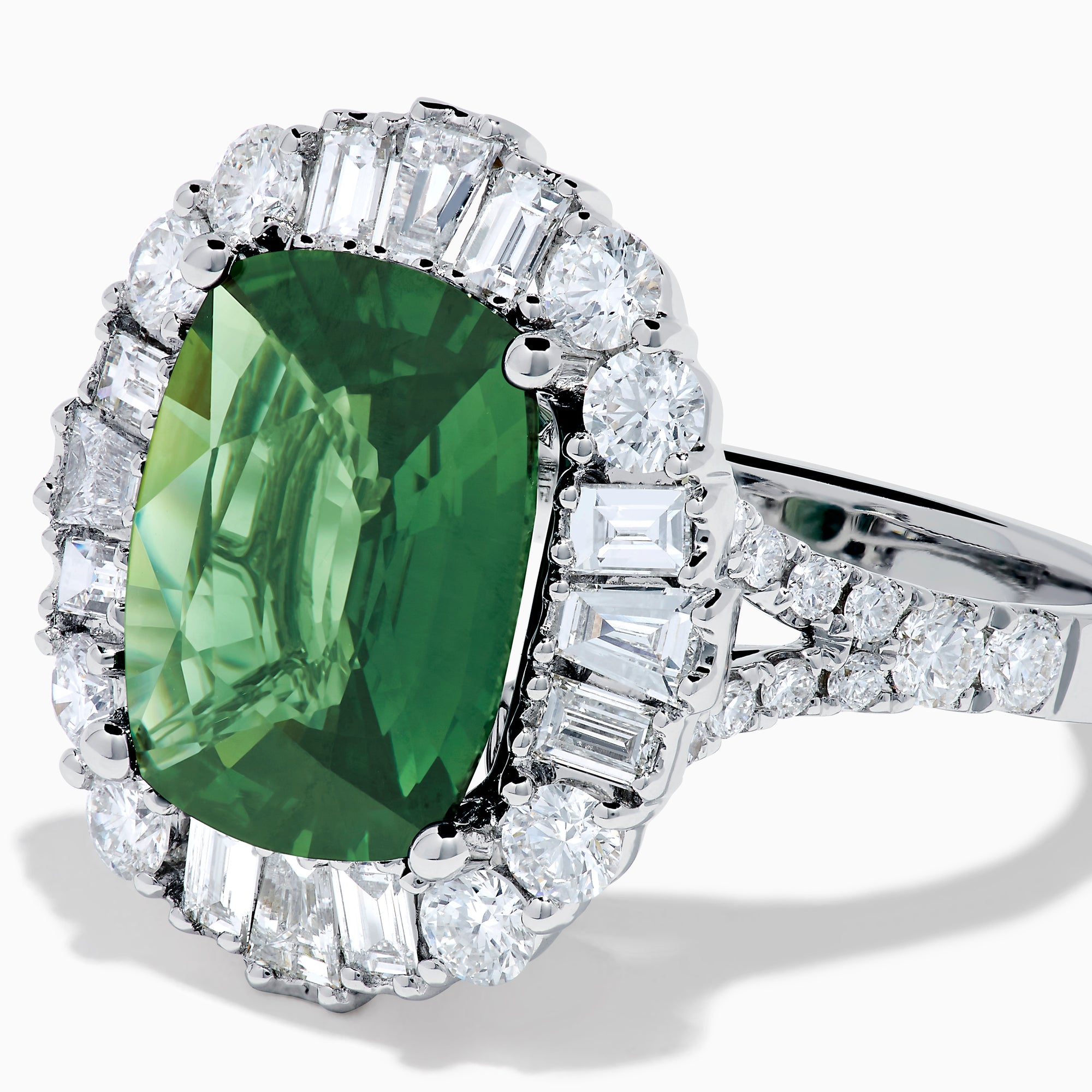 The 18K White Gold Green Sapphire and Diamond Halo Ring showcases a large oval green sapphire, surrounded by round and rectangular white diamonds, set on an elegant white gold band and displayed on a white background.