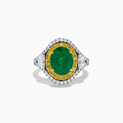 The 18K Gold Emerald, Yellow and White Diamond Double Halo Ring features a large oval green emerald center, surrounded by double halos of yellow and white diamonds with side diamonds on an 18K gold band. Set against a plain white background.