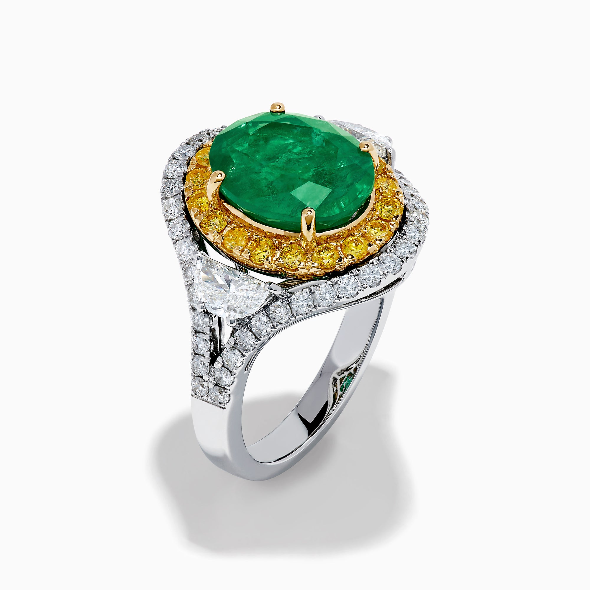 The 18K Gold Emerald, Yellow and White Diamond Double Halo Ring features a large oval green emerald center, surrounded by double halos of yellow and white diamonds with side diamonds on an 18K gold band. Set against a plain white background.