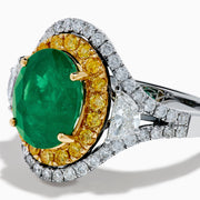 The 18K Gold Emerald, Yellow and White Diamond Double Halo Ring features a large oval green emerald center, surrounded by double halos of yellow and white diamonds with side diamonds on an 18K gold band. Set against a plain white background.