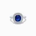 The 18K White Gold Blue Sapphire and Diamond Double Halo Ring showcases a large cushion-cut blue sapphire encircled by two rows of diamonds, elegantly displayed on a plain white background.