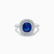 The 18K White Gold Blue Sapphire and Diamond Double Halo Ring showcases a large cushion-cut blue sapphire encircled by two rows of diamonds, elegantly displayed on a plain white background.