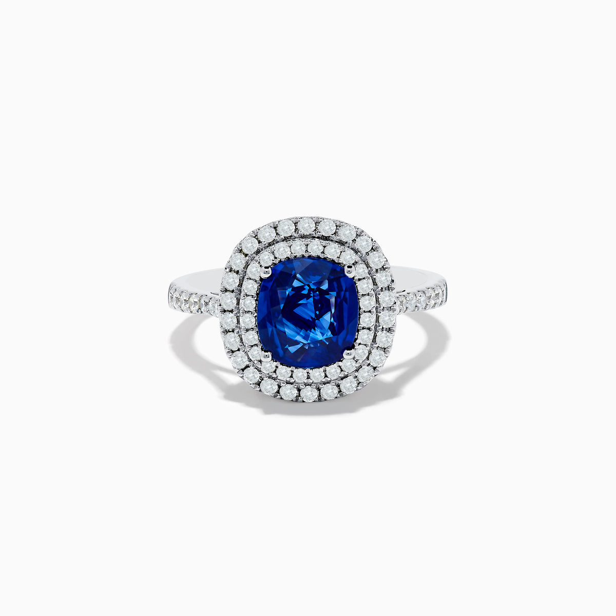 The 18K White Gold Blue Sapphire and Diamond Double Halo Ring showcases a large cushion-cut blue sapphire encircled by two rows of diamonds, elegantly displayed on a plain white background.