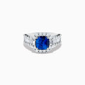 The 18K White Gold GIA Certified Cushion Cut Blue Sapphire and Diamond Ring features a striking cushion cut blue sapphire center, round diamond halo, and baguette-cut diamonds on the band, shown against a white background.