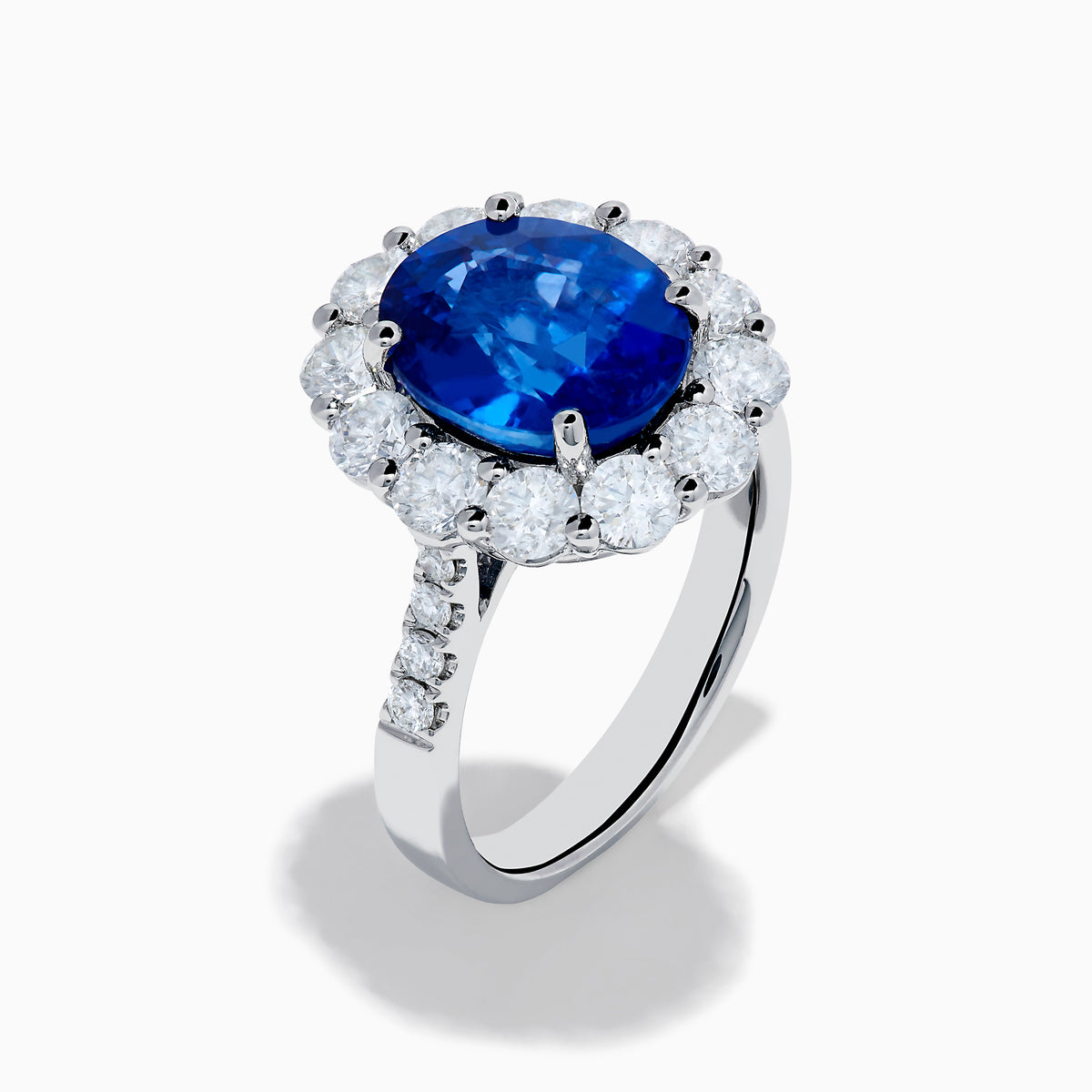 The 18K White Gold Blue Sapphire and Diamond Halo Ring features a large oval blue sapphire surrounded by a halo of round diamonds, showcased on a white background.