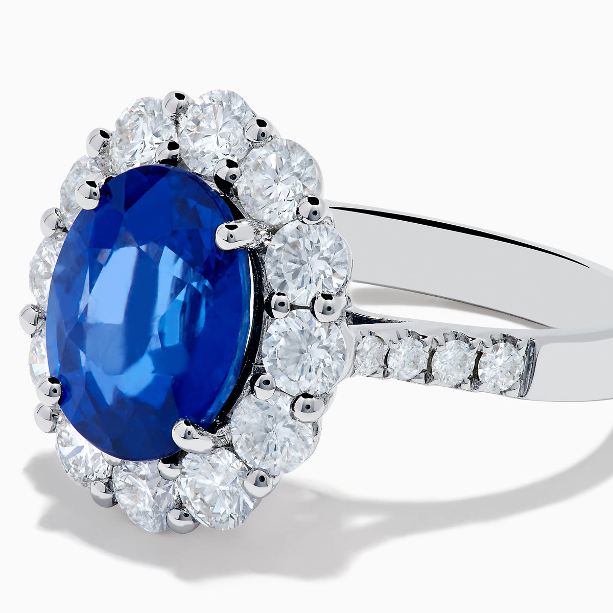 The 18K White Gold Blue Sapphire and Diamond Halo Ring features a large oval blue sapphire surrounded by a halo of round diamonds, showcased on a white background.