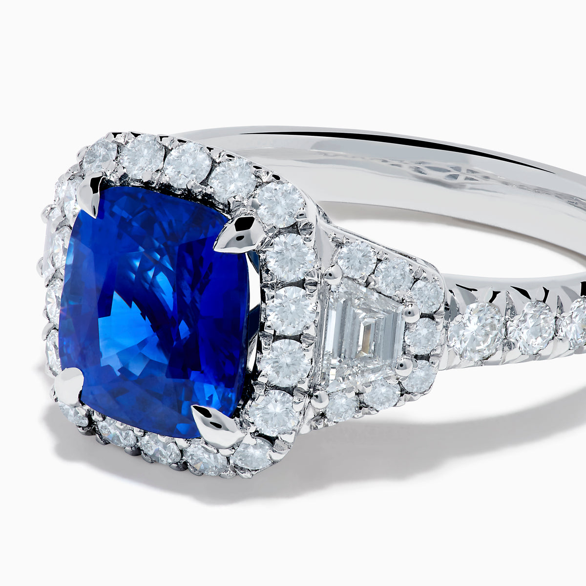 The Royale Bleu Platinum Blue Sapphire Halo Ring showcases a large cushion-cut sapphire surrounded by sparkling diamonds on a platinum band, set against a white background for a timeless, elegant look.