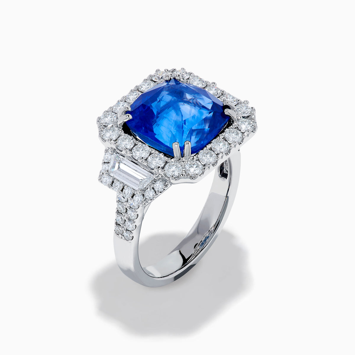 The Royale Bleu 18K White Gold Blue Sapphire and Diamond Halo Ring showcases a cushion-cut blue sapphire center with a double diamond halo and diamond side accents, elegantly set against a white background.