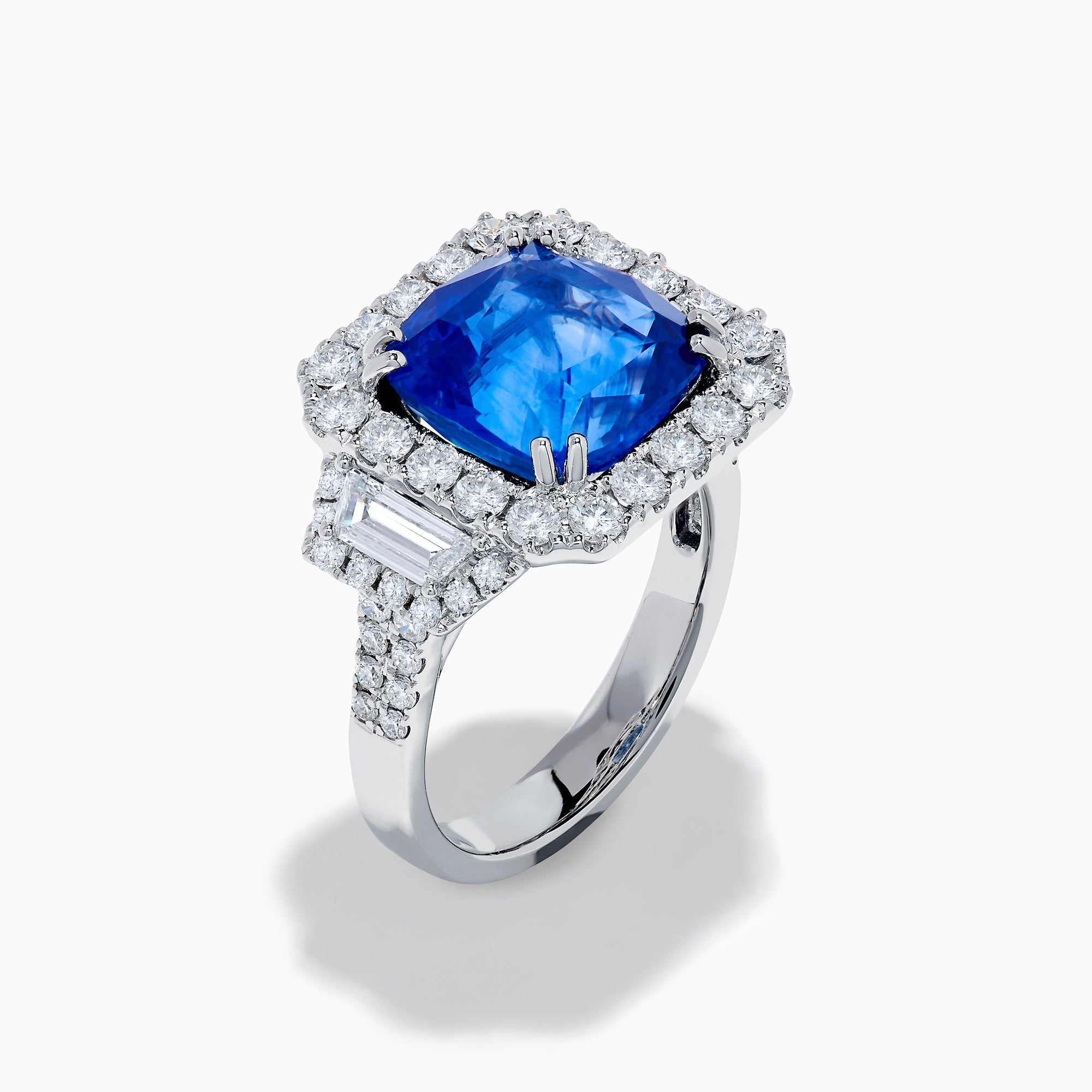 The Royale Bleu 18K White Gold Blue Sapphire and Diamond Halo Ring showcases a cushion-cut blue sapphire center with a double diamond halo and diamond side accents, elegantly set against a white background.