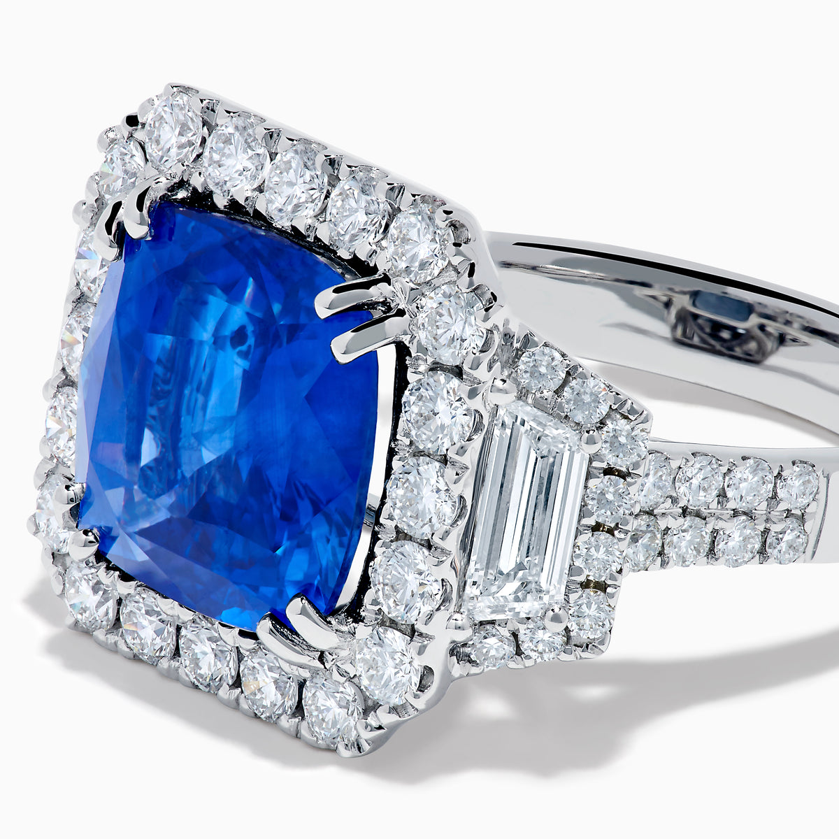 The Royale Bleu 18K White Gold Blue Sapphire and Diamond Halo Ring showcases a cushion-cut blue sapphire center with a double diamond halo and diamond side accents, elegantly set against a white background.