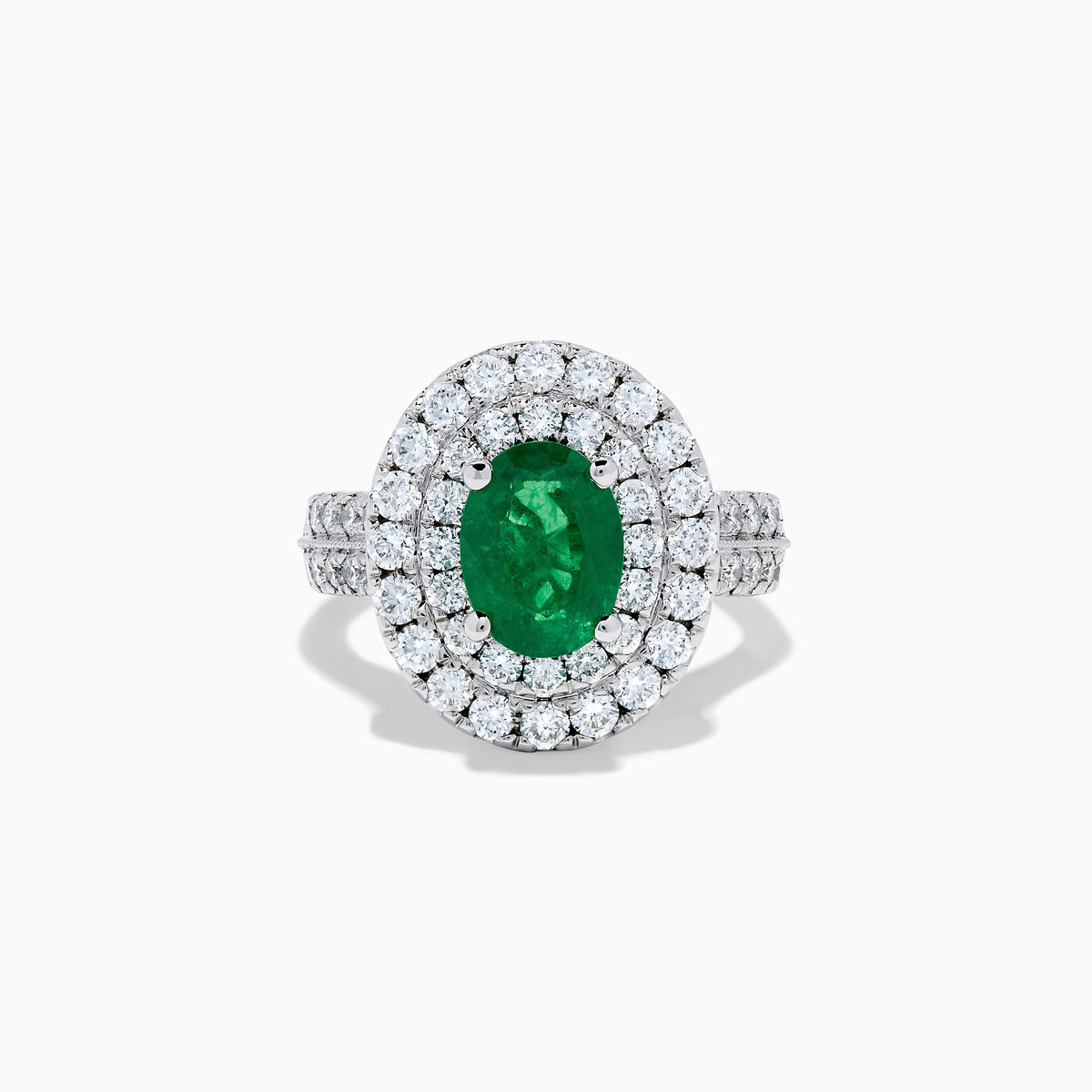 The Brasilica 18K White Gold Emerald and Diamond Double Halo Ring features an oval green emerald encircled by two rows of diamonds and delicate side stones, all set on a white gold band against a white background.